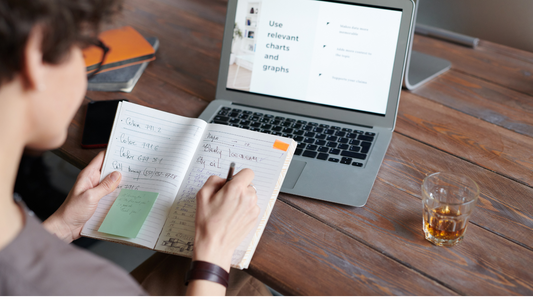 a person writing on a notebook or diary with an open laptop in front of them