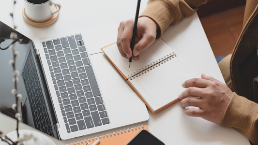 a person writing on a notebook in front of an open laptop